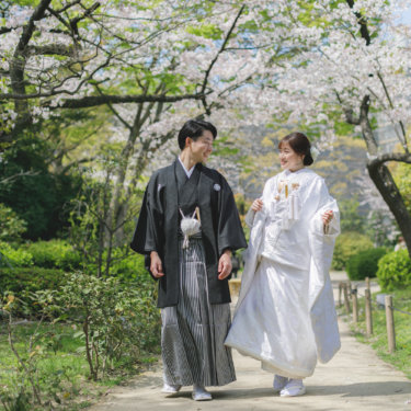 桜並木の道を並んで歩く黒紋付袴の新郎と白無垢の花嫁を引きでとらえた一枚。桜で撮る春の和装前撮り