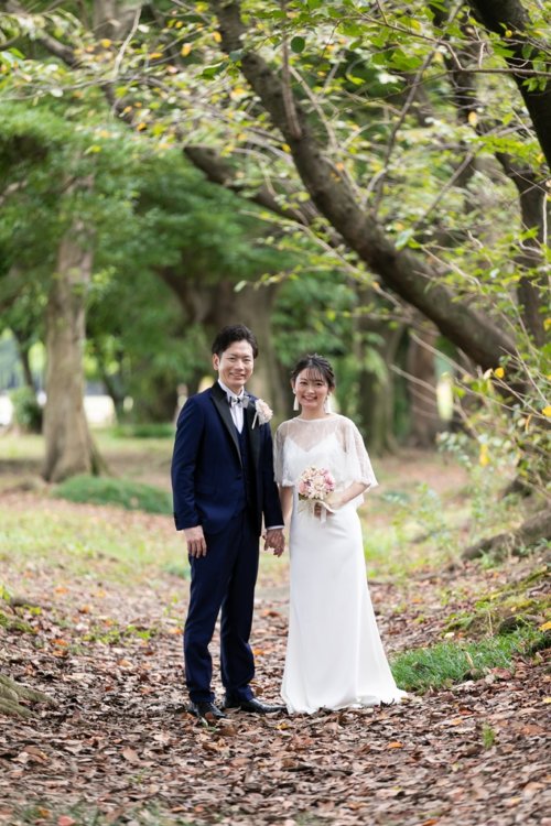 落ち葉の敷きつめられた並木道で手をつなぐ新郎新婦。ネイビータキシードとレースケープを重ねた白いドレスの洋装ロケーションフォト