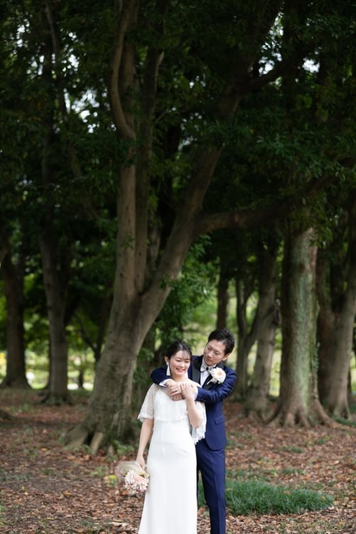 大きな木々の下で花嫁を後ろからそっと抱き寄せる新郎。落ち葉の絨毯が広がる木立での自然体なフォトウェディング