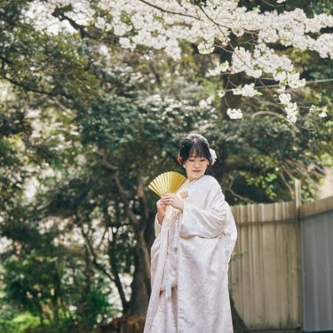 桜の木の下で金色の扇子を持って微笑む白い色打掛の花嫁を引きでとらえた一枚。春の桜で撮る和装前撮り