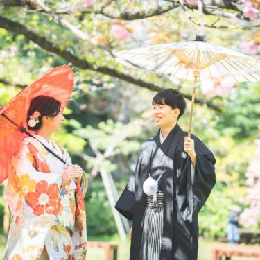 桜の下で赤い和傘と白い和傘を差して見つめ合う花柄の色打掛の花嫁と黒紋付袴の新郎。春の桜で撮る和装ロケーションフォト