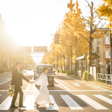 黄葉のイチョウ並木の横断歩道を手をつないで渡る黒いタキシードの新郎とウェディングドレスの花嫁。秋の街中で撮る洋装前撮り