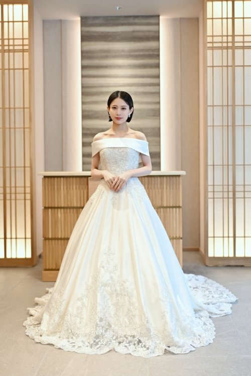 木の格子の壁の前でオフショルダーのサテンのウェディングドレスを着て手を重ねる花嫁を縦位置でとらえた一枚。東京駅店のスタジオで撮る洋装前撮りのドレス写真