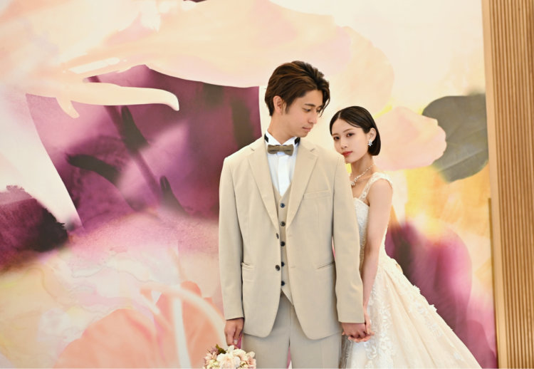 紫と桃色の花のアートウォールの前で手をつなぐベージュのスーツの新郎とレースのウェディングドレスの花嫁。東京駅店のスタジオで撮る洋装前撮り