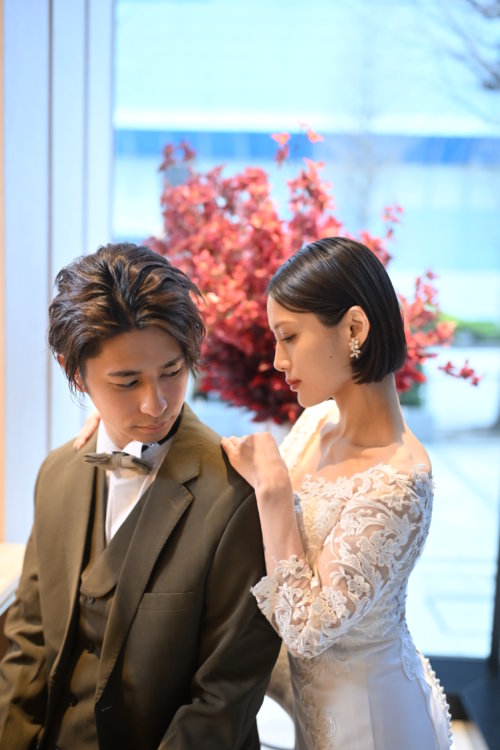赤い葉のオブジェを背に新郎の肩に手を添えるレースの袖のウェディングドレスの花嫁とオリーブのスーツの新郎を縦位置でとらえた一枚。東京駅店のスタジオで撮る洋装前撮り