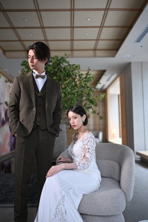 緑の木を背にソファに座るレースの袖のウェディングドレスの花嫁と、隣に立つオリーブのスーツの新郎を縦位置でとらえた一枚。東京駅店のラウンジで撮る洋装前撮り