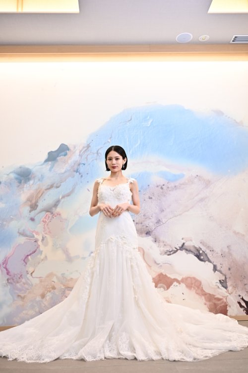 水彩模様のアートウォールの前でレースのマーメイドドレスを着て手を重ねる花嫁を縦位置でとらえた一枚。洋装前撮りで選べるウェディングドレスの衣装写真