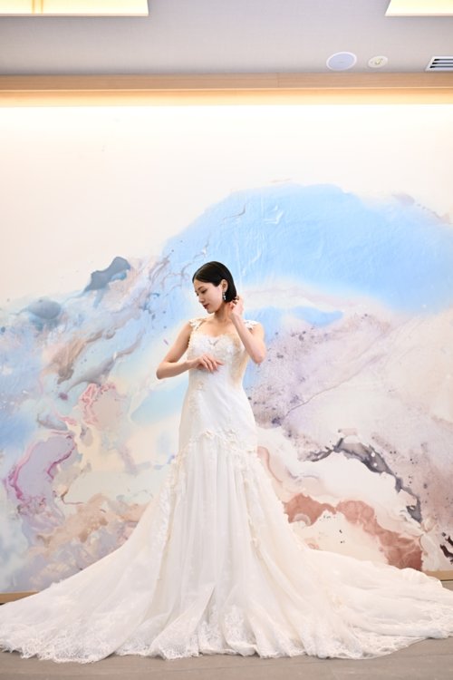 水彩模様のアートウォールの前でレースのマーメイドドレスを着て髪に手を添える花嫁を縦位置でとらえた一枚。東京駅店のスタジオで撮る洋装前撮りのドレス写真