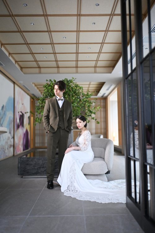 緑の木の飾られたラウンジのソファに座るレースの袖のウェディングドレスの花嫁と立つオリーブのスーツの新郎を縦位置でとらえた一枚。東京駅店のラウンジで撮る洋装前撮り