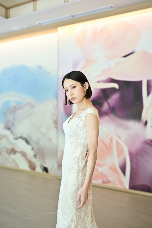 紫と桃色の花のアートウォールの前でVネックのレースのウェディングドレスを着て立つ花嫁を縦位置でとらえた一枚。東京駅店のスタジオで撮る洋装前撮りのドレス写真