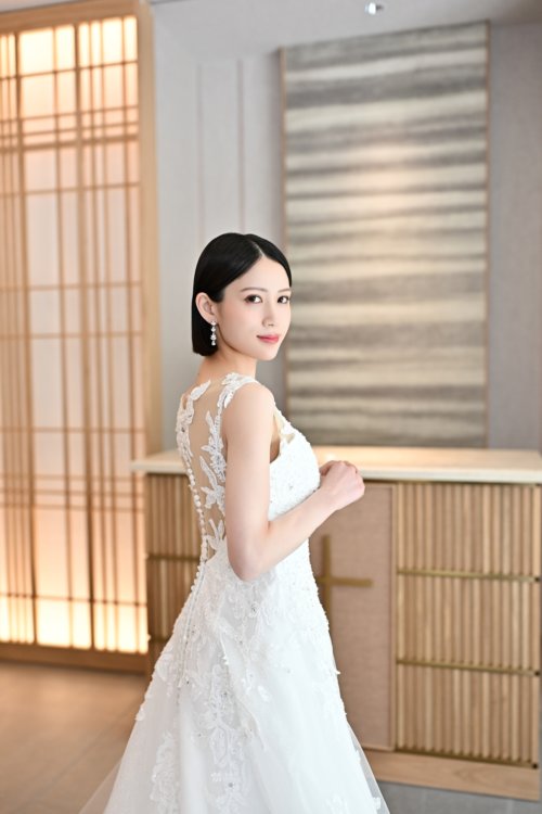 木の格子の壁の前で背中の透けるレースのウェディングドレスを着て振り返る花嫁を縦位置でとらえた一枚。東京駅店のスタジオで撮る洋装前撮りのドレス写真