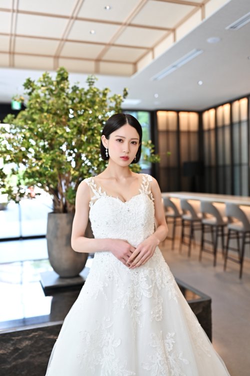 緑の木を背にレースのAラインのウェディングドレスを着て立つ花嫁を縦位置でとらえた一枚。東京駅店のラウンジで撮る洋装前撮りのドレス写真