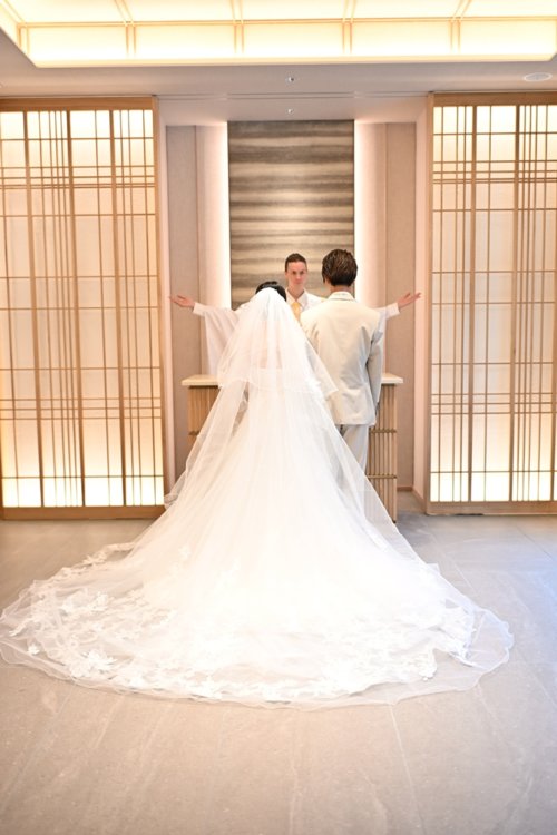 祭壇で両手を広げる牧師の前に立つベージュのスーツの新郎とベールのウェディングドレスの花嫁を後ろから縦位置でとらえた一枚。東京駅店のスタジオで撮るチャペルフォト