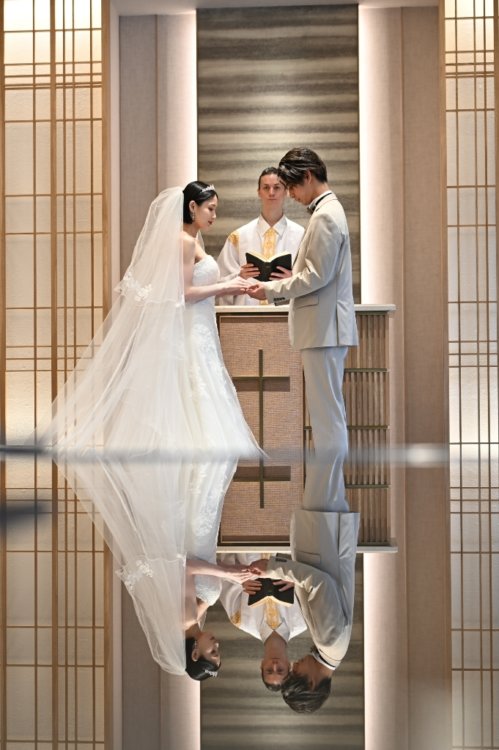 祭壇で牧師と向かい合うベージュのスーツの新郎とベールのウェディングドレスの花嫁と、床に映る姿を縦位置でとらえた一枚。東京駅店のスタジオで撮るリフレクションのチャペルフォト