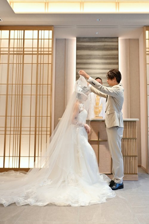 祭壇の前で花嫁のベールを上げるベージュのスーツの新郎とウェディングドレスの花嫁を縦位置でとらえた一枚。東京駅店のスタジオで撮るチャペルフォトのベールアップ