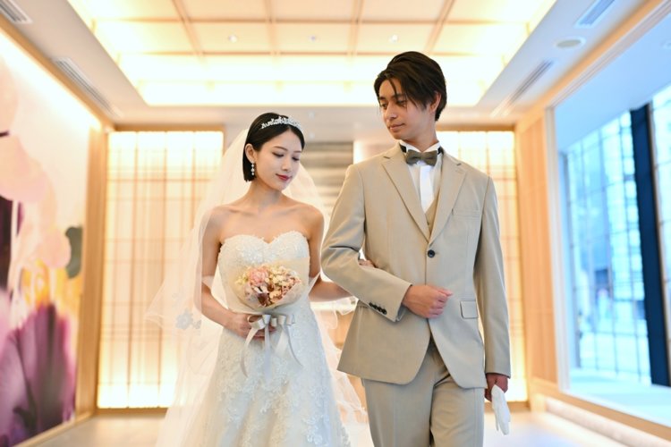 木の格子の壁の前でティアラとベールを付けてブーケを持ち、ベージュのスーツの新郎と腕を組むウェディングドレスの花嫁。東京駅店のスタジオで撮るチャペルフォト