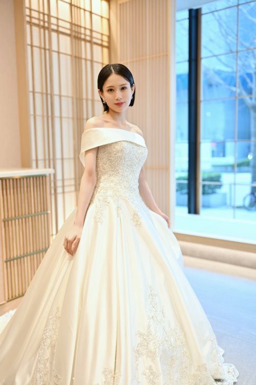 街路樹を望む窓辺でオフショルダーのサテンのウェディングドレスの裾を持って立つ花嫁を縦位置でとらえた一枚。東京駅店のスタジオで撮る洋装前撮りのドレス写真