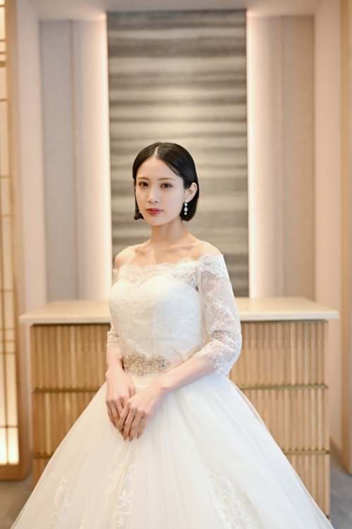 木の格子の壁の前でレースの袖のオフショルダーのウェディングドレスを着て手を重ねる花嫁を縦位置でとらえた一枚。東京駅店のスタジオで撮る洋装前撮りのドレス写真