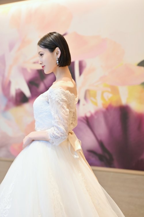 紫と桃色の花のアートウォールの前でレースの袖のウェディングドレスを着て横を向く花嫁を縦位置でとらえた一枚。洋装前撮りで選べる袖付きウェディングドレスの衣装写真