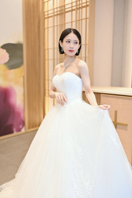 紫と桃色の花のアートウォールの前でビスチェのチュールのウェディングドレスの裾を持つ花嫁を縦位置でとらえた一枚。洋装前撮りで選べるウェディングドレスの衣装写真