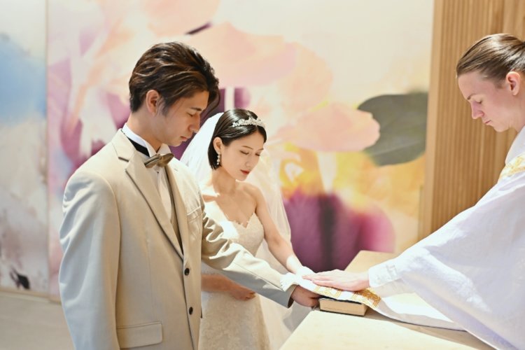 アートウォールの前で聖書に手を重ねるベージュのスーツの新郎とベールのウェディングドレスの花嫁と牧師。東京駅店のスタジオで撮るチャペルフォトの誓いの一場面