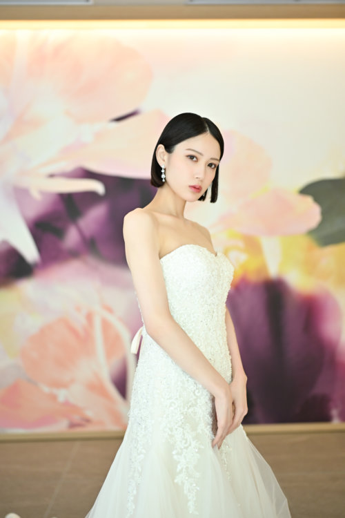 紫と桃色の花のアートウォールの前でレースのウェディングドレスを着て立つ花嫁を縦位置でとらえた一枚。東京駅店のスタジオで撮る洋装前撮りのドレス写真