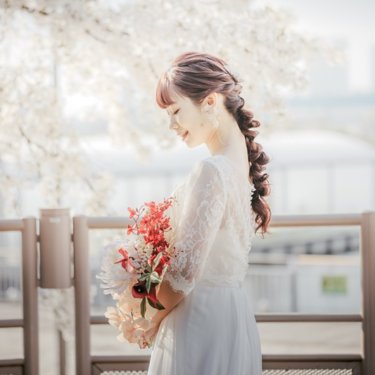 満開の桜の下で赤い花のブーケを持って目を伏せる編み込みヘアのレースのウェディングドレスの花嫁。春の桜で撮る洋装前撮り