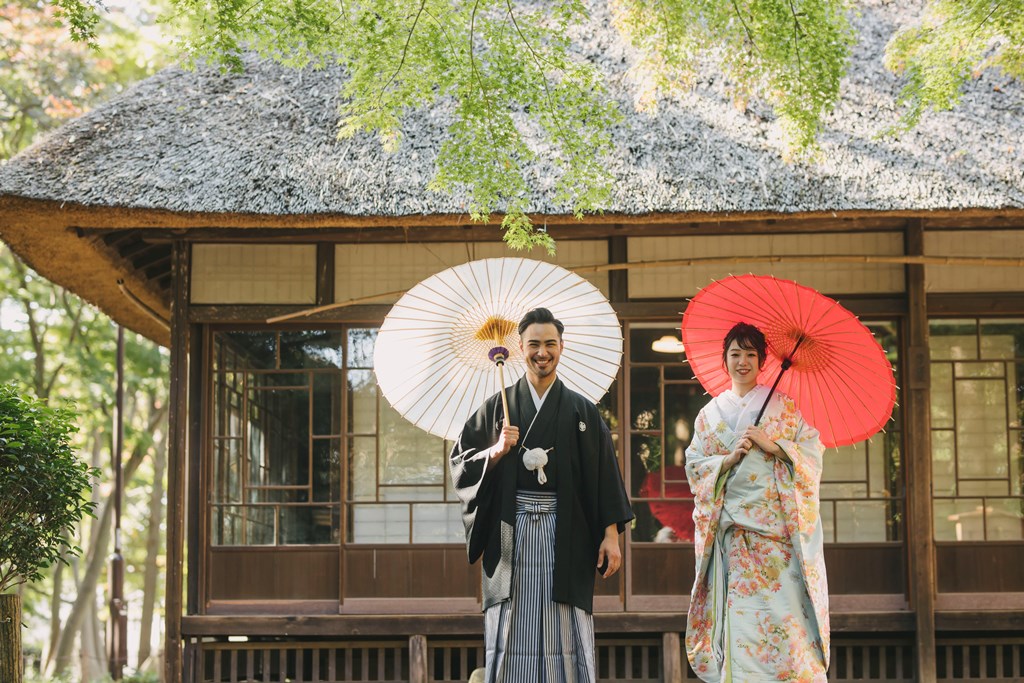 蘆花恒春園の茅葺きの日本家屋を背に、白と赤の和傘を手にした紋付袴の新郎と色打掛の花嫁。和装ロケーション前撮り