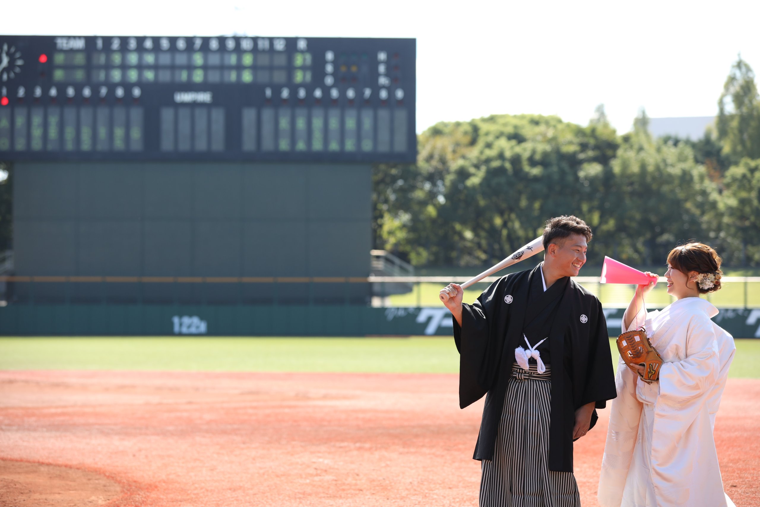 野球場のスコアボードを背景に、バットを肩に担ぎ笑い合う黒紋付袴姿の新郎とピンクのメガホンを掲げる白無垢姿の花嫁。