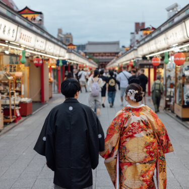 仲見世通りの石畳を手をつないで歩く新郎新婦の後ろ姿。花嫁のかんざしと色打掛の背中の柄が映える浅草の前撮り写真