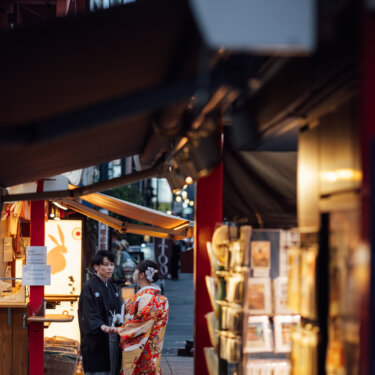 土産物店の軒先越しに、店先で向き合う新郎新婦をのぞき見るように撮った和装の結婚写真。前ボケを活かしたおしゃれな構図