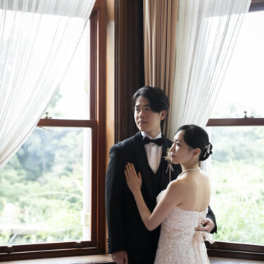 窓辺で新郎に寄り添い、横顔を見せる花嫁。パールのネックレスとまとめ髪が上品なクラシカルな前撮り写真