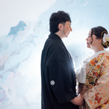 紫の花のアートウォールの前に並ぶ黒紋付袴の新郎と白無垢の花嫁を縦位置でとらえた一枚。東京駅店のスタジオで撮る和装前撮り