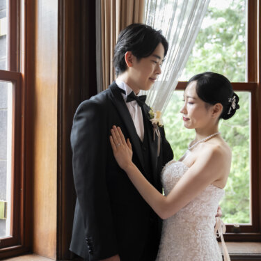 木枠の窓の前で、新郎の胸元に手を当てて見上げる花嫁。やわらかな自然光に包まれた洋館のウェディングフォト