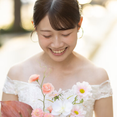ピンクのガーベラやアンスリウムのブーケを胸元に、うつむいてほほえむ花嫁のアップ。やわらかな光のウェディングフォト