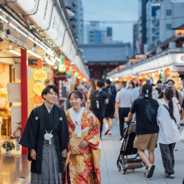 夕暮れの仲見世通りを笑顔で歩く黒紋付袴の新郎と色打掛の花嫁。観光客が行き交う浅草の賑わいを残した自然体の和装前撮り