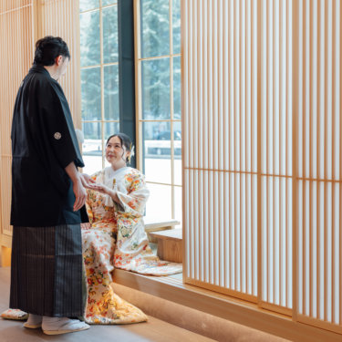 青いアートウォールの前で手をつなぐ黒紋付袴の新郎と白無垢の花嫁を縦位置でとらえた一枚。東京駅店のスタジオで撮る和装スタジオフォト