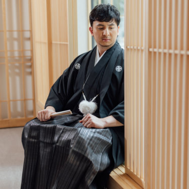 障子の前で花嫁を見て笑う黒紋付袴の新郎と、髪飾りを付けた白無垢の花嫁の後ろ姿。東京駅店のスタジオで撮る和装前撮り
