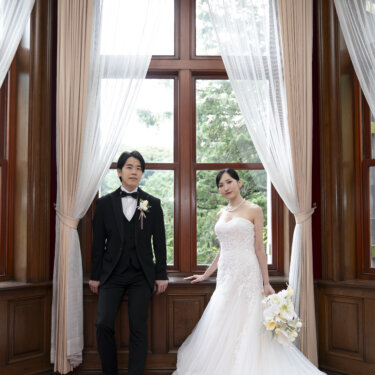 木枠の出窓の前に並んで立つタキシードの新郎とウェディングドレスの花嫁。旧古河邸の重厚な内装を活かした前撮り写真