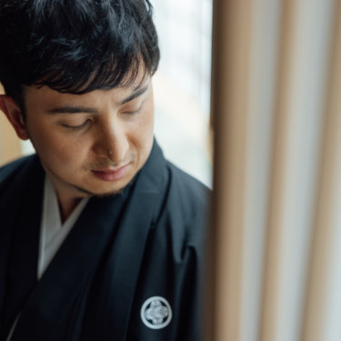 縦格子の障子の前で白い和傘とピンクの和傘を差して向かい合う黒紋付袴の新郎と白無垢の花嫁。和傘を使った和装前撮り