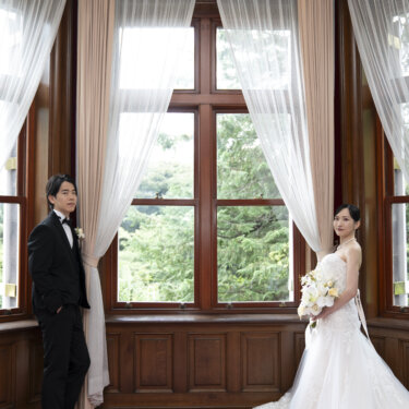 旧古河邸にて、大きな窓の両端に離れて立つ新郎と花嫁を、窓越しの緑とともに収めた洋館でのフォトウェディング
