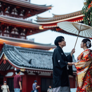 浅草寺の五重塔と本堂の屋根を背に、白い和傘をかざして手をつなぐ黒紋付袴の新郎と色打掛の花嫁。王道の浅草和装フォトウェディング