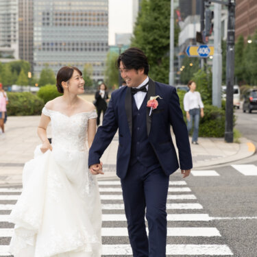 緑のある広場の横断歩道を、手をつないで笑顔で歩くタキシードの新郎とウェディングドレスの花嫁。東京駅周辺で撮る自然体の結婚写真