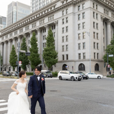 高層ビルと石造りの建物を見上げる構図で、横断歩道を渡る新郎新婦を写した丸の内の街ロケ前撮り