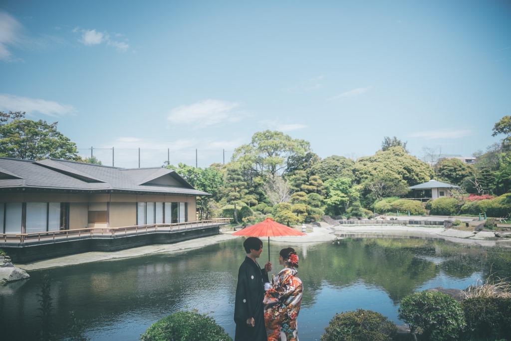 茅葺屋根の東屋で見つめ合う黒紋付袴姿の新郎と白無垢姿の花嫁。手前の緑葉が幻想的にボケた一枚。