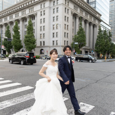円柱の並ぶクラシカルなビルの前の横断歩道を、ドレスの裾を持って歩く花嫁と新郎。丸の内の街並みで撮るフォトウェディング