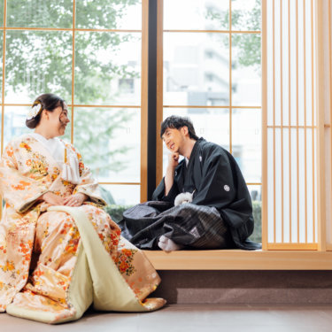 縦格子の障子の前で白い和傘の下に寄り添う黒紋付袴の新郎と白無垢の花嫁。東京駅店のスタジオで撮る和装前撮り