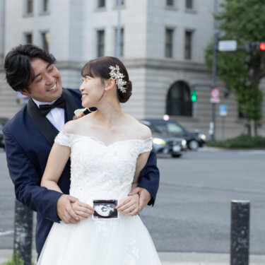 重厚な石造りのビルが並ぶ丸の内の交差点で、後ろから花嫁を抱き寄せエコー写真を見せる新郎。マタニティ前撮りのほほえましい一枚