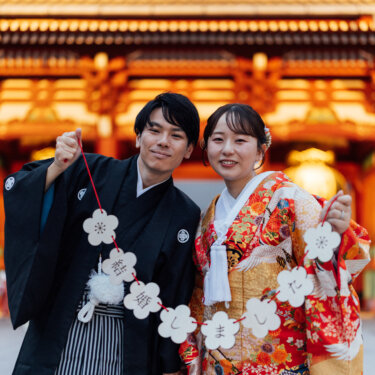 梅の形のガーランドを掲げて笑顔を見せる羽織袴の新郎と色打掛の花嫁。浅草寺の朱色の門の前で撮った結婚報告にも使える前撮り写真