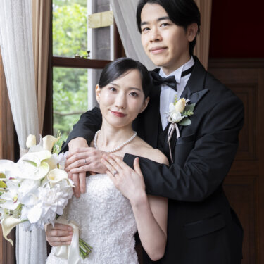 窓辺で花嫁を後ろから抱きしめ、肩に手を重ねる新郎。結婚指輪がのぞく洋装の結婚写真